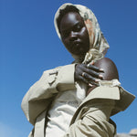 A model wearing a beige trench coat over a white top and trousers, paired with a patterned beige and orange headscarf, set against a bright blue sky and rocky background.