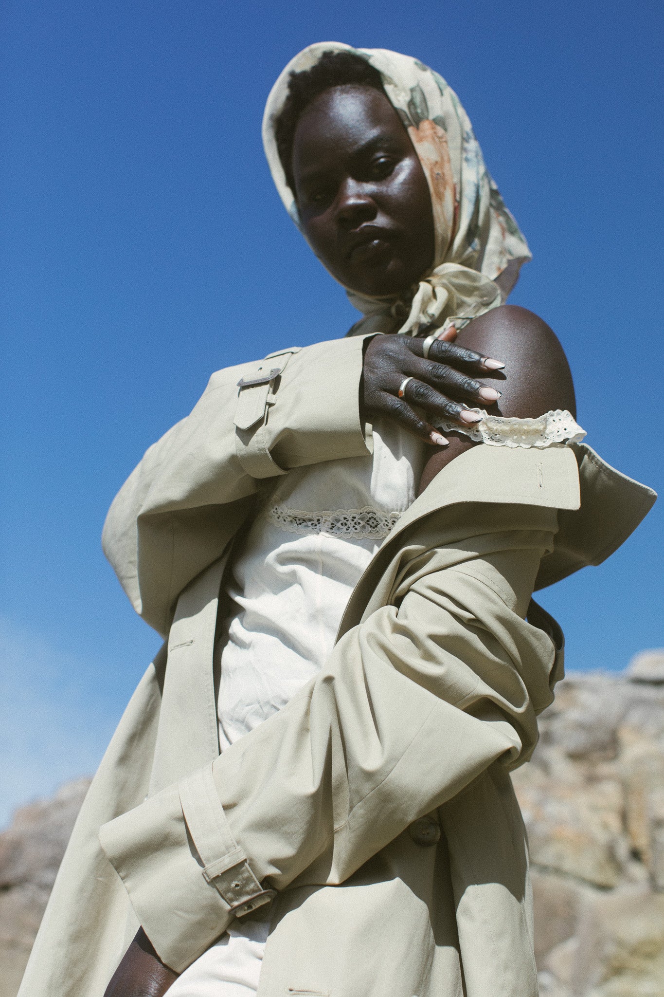 A model wearing a beige trench coat over a white top and trousers, paired with a patterned beige and orange headscarf, set against a bright blue sky and rocky background.