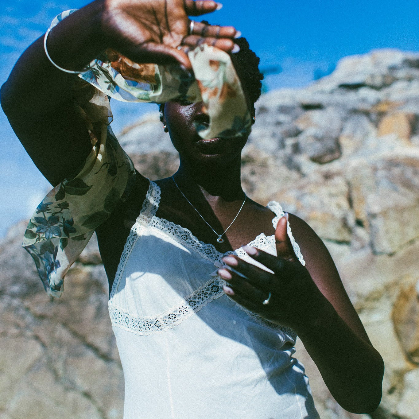 A woman outdoors wearing a white lace-trimmed dress, holding a patterned scarf in front of her face, with rocky cliffs and a bright blue sky in the background.