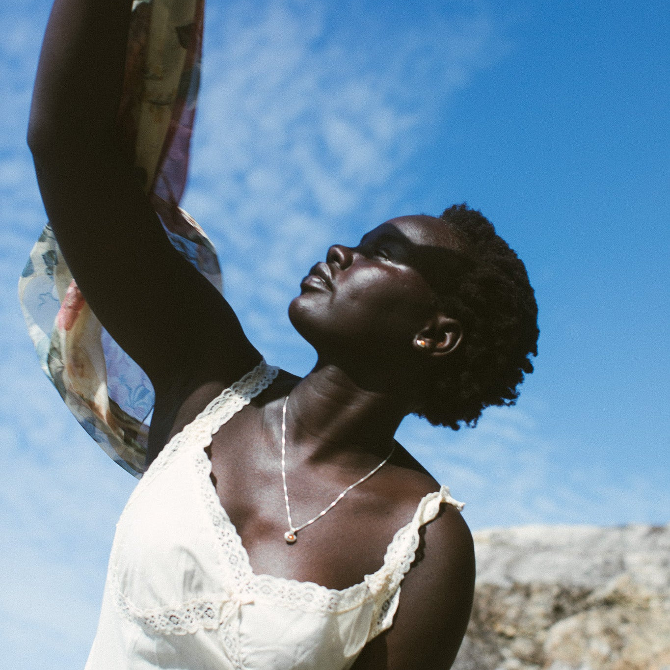 A woman outdoors under a bright blue sky, wearing a white lace-trim camisole and holding a patterned scarf above her head. She is also wearing the sunstone siren stud earrings. 