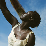A woman wearing a cream lace-trim camisole and gold jewelry looks upward against a bright blue sky, holding a patterned scarf.