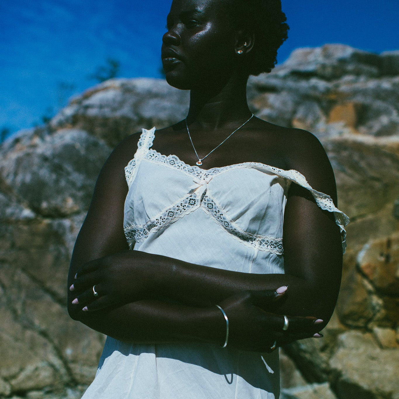 A woman wearing a white off-the-shoulder lace-trim dress stands outdoors against a rocky background under a blue sky.