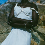 A woman wearing a white off-the-shoulder lace-trim dress stands outdoors against a rocky background under a blue sky.