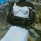A woman wearing a white off-the-shoulder lace-trim dress stands outdoors against a rocky background under a blue sky.