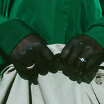 Close-up of a person's hands buttoning a light beige jacket or pants, wearing a vibrant green long-sleeve shirt and silver rings.