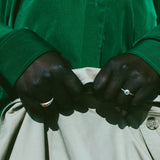Close-up of a person's hands buttoning a light beige jacket or pants, wearing a vibrant green long-sleeve shirt and silver rings.