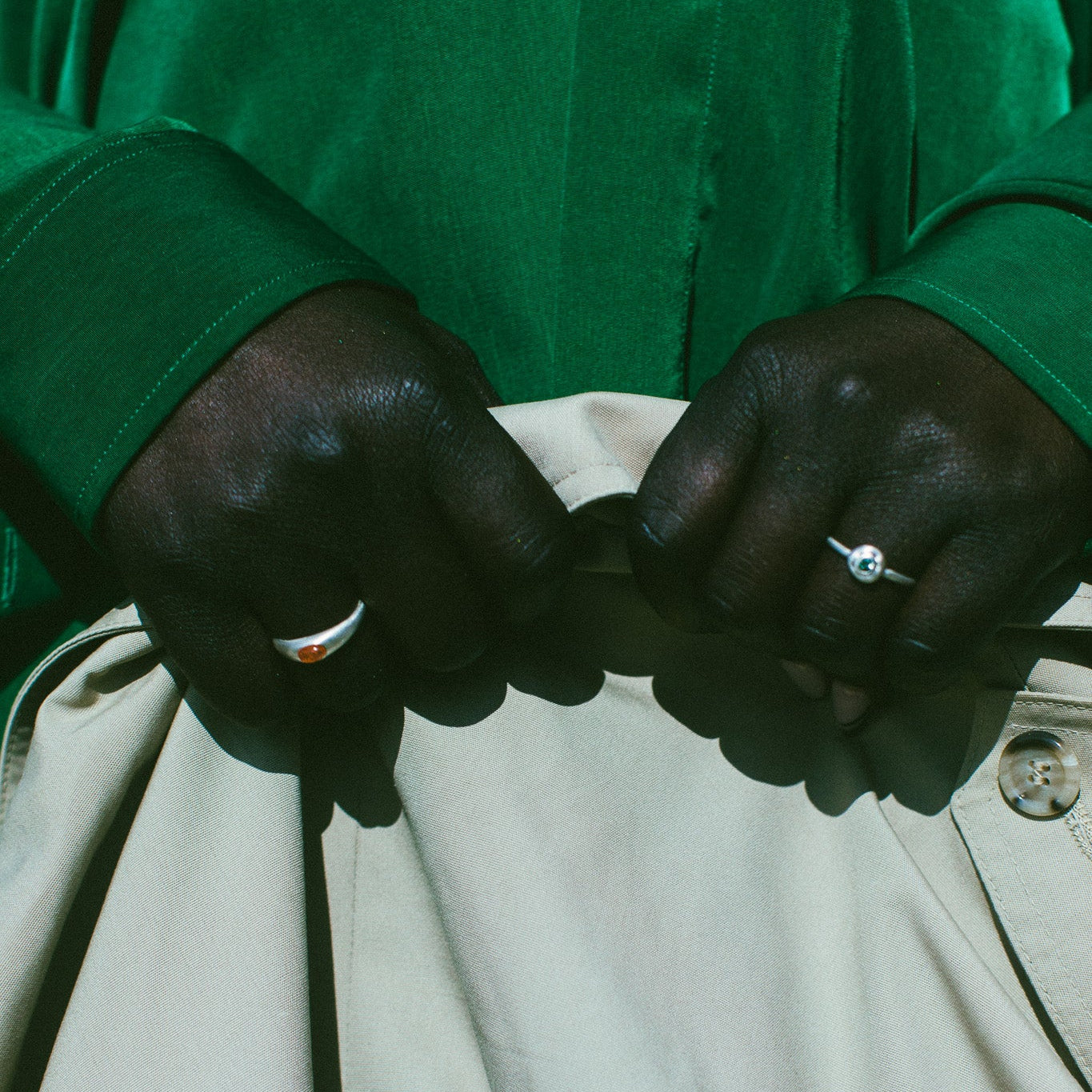 Close-up of a person's hands buttoning a light beige jacket or pants, wearing a vibrant green long-sleeve shirt and silver rings.