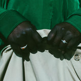 Close-up of a person's hands buttoning a light beige jacket or pants, wearing a vibrant green long-sleeve shirt and silver rings.