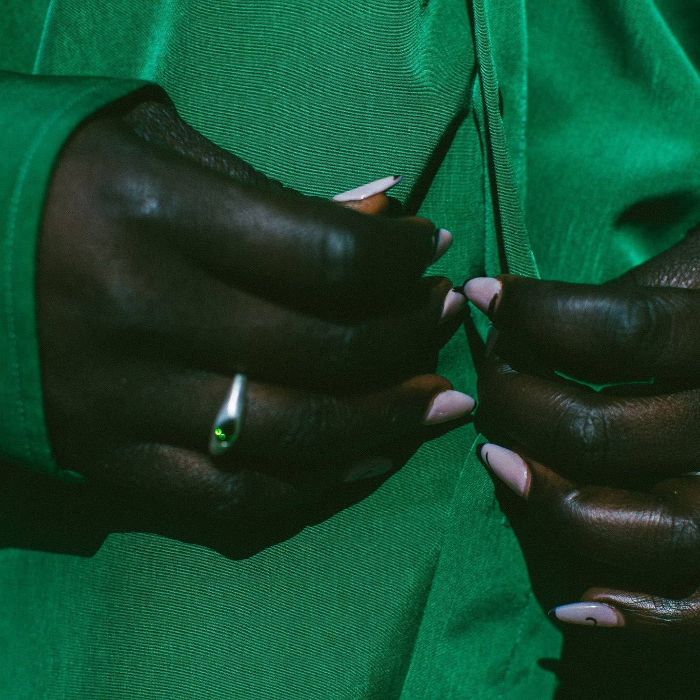 Close-up of a person wearing a vibrant green satin-like top or dress, with their hands buttoning the front. The person is wearing a silver ring with a green stone on one finger.