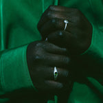 A close-up of a person's hands wearing two silver rings, one with a small round clear gemstone and the other with a small oval green gemstone, set against a dark green satin jacket.