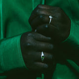 A close-up of a person's hands wearing two silver rings, one with a small round clear gemstone and the other with a small oval green gemstone, set against a dark green satin jacket.