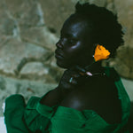 A woman in profile wearing a dark green draped garment, with a bright yellow flower tucked behind her ear, set against a textured stone wall.