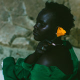 A woman in profile wearing a dark green draped garment, with a bright yellow flower tucked behind her ear, set against a textured stone wall.