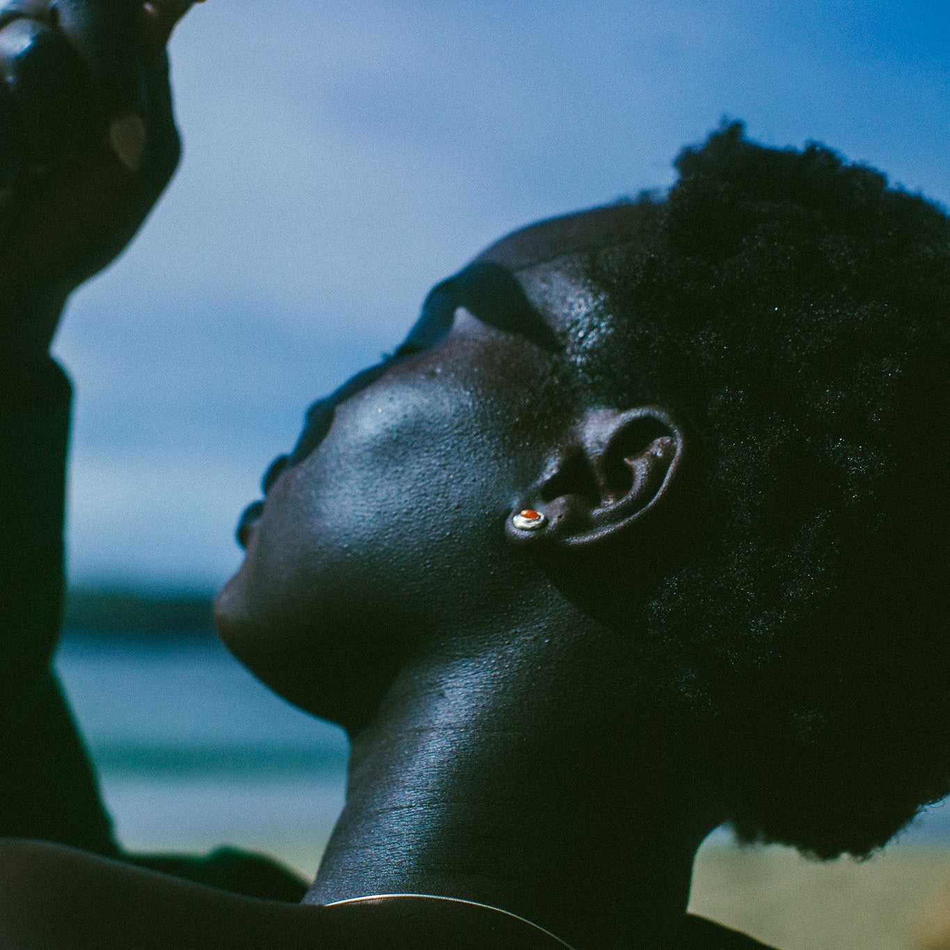 A close-up lifestyle shot of a person with dark skin looking upward. They are wearing a small silver stud featuring an oval sunstone, with a blue sky and blurred outdoor background.