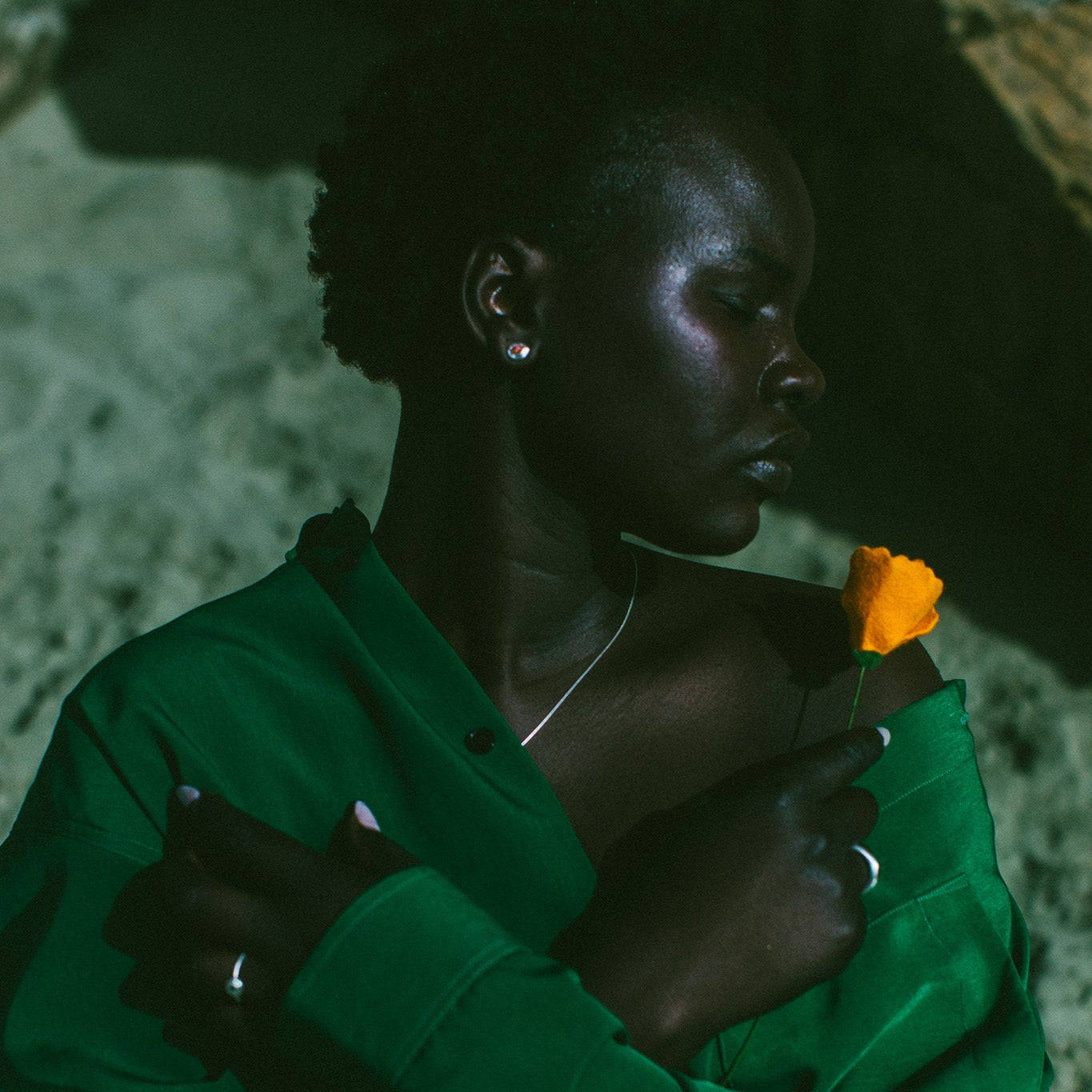 A woman in a dark green satin-like button-up shirt wearing silver earrings with an orange stone in the centre, 
holding an orange flower, set against a moody, shadowed stone background.