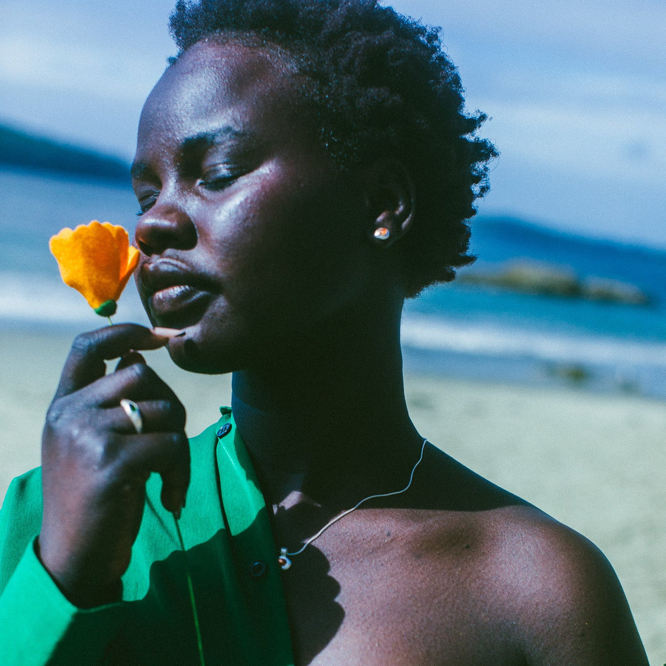 A woman on a beach wearing a bright green off-the-shoulder shirt, and silver earrings with an orange stone in the centre, holding a yellow flower near her face. The background shows sand and the ocean.
