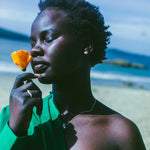 A woman on a beach wearing a bright green off-the-shoulder shirt, and silver earrings with an orange stone in the centre, holding a yellow flower near her face. The background shows sand and the ocean.