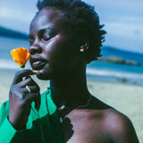 A woman on a beach wearing a bright green off-the-shoulder shirt, and silver earrings with an orange stone in the centre, holding a yellow flower near her face. The background shows sand and the ocean.