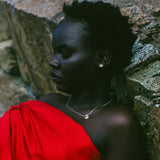 A woman with short dark hair leans against a stone wall, wearing a bright red draped garment and a delicate silver necklace with a small oval pendant.