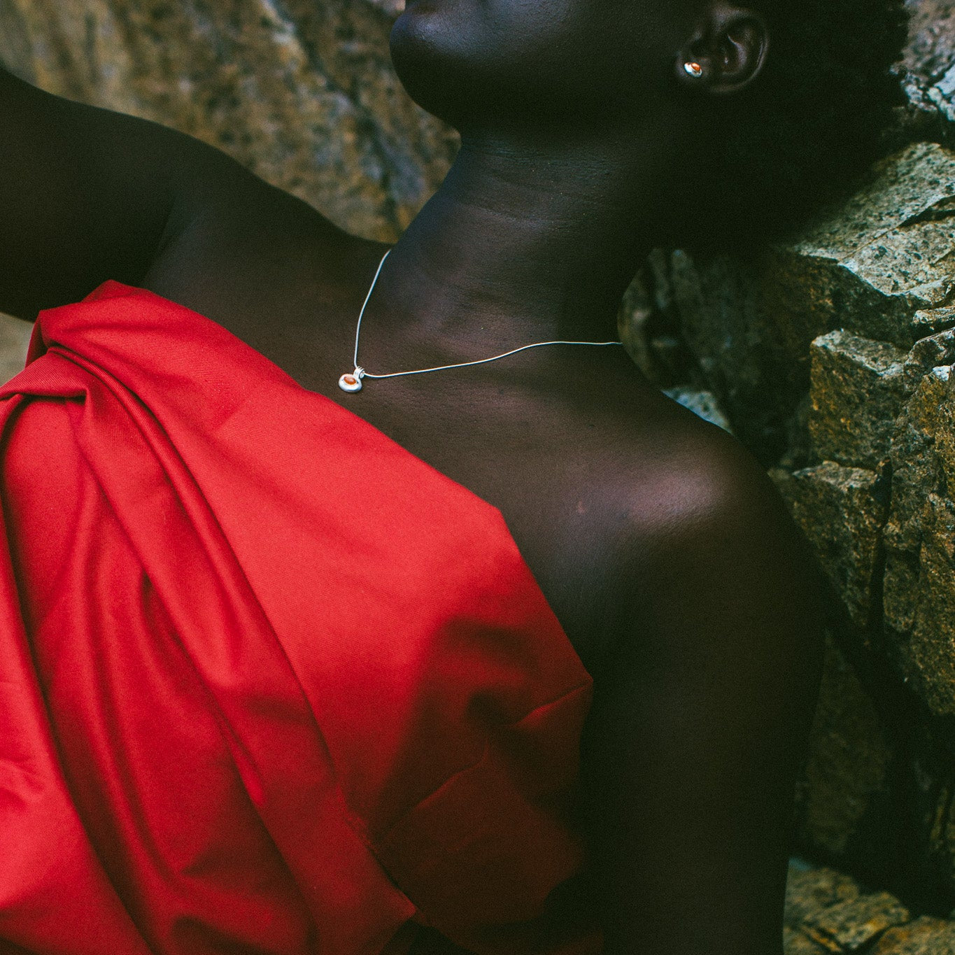A close-up lifestyle shot of a model wearing a vibrant red draped dress or wrap, resting against a textured stone wall. The model has dark skin and is wearing a delicate silver necklace.