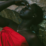 A woman reclining against a rocky surface, wearing a bright red draped garment and a delicate silver necklace.
