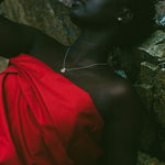 A woman reclining against a rocky surface, wearing a bright red draped garment and a delicate silver necklace.