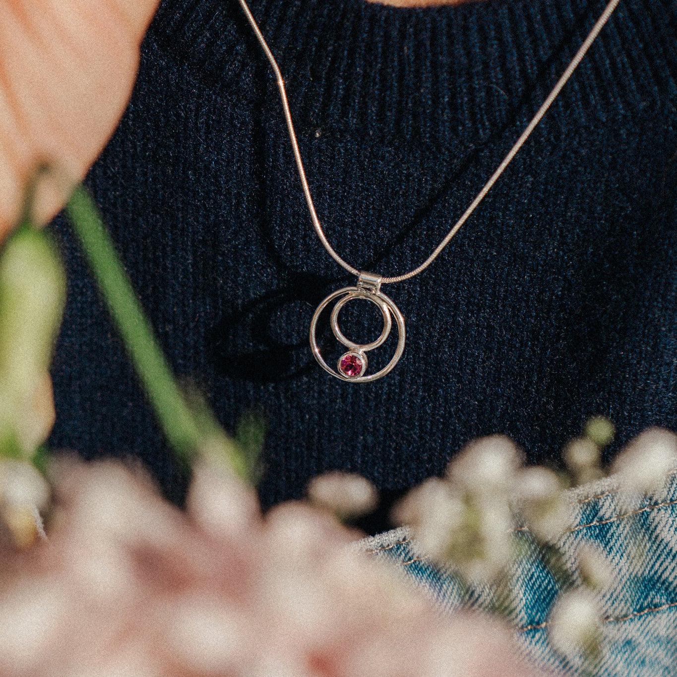 Person wearing a necklace with a circular pendant featuring a red gemstone, surrounded by flowers.