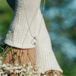 Person wearing a necklace with a pendant, standing among white flowers with a blurred natural background