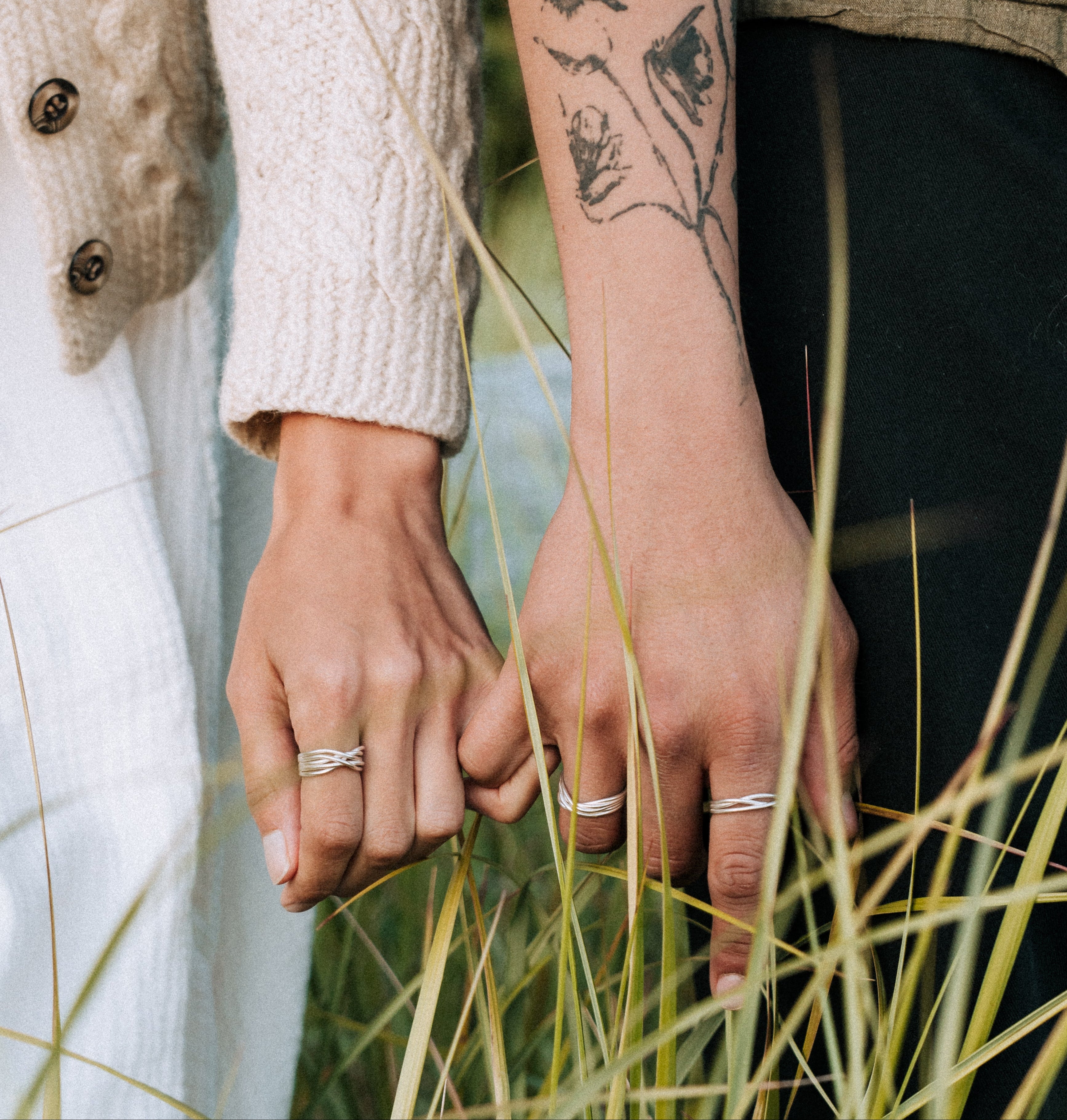 Two people holding hands with rings, standing in tall grass.