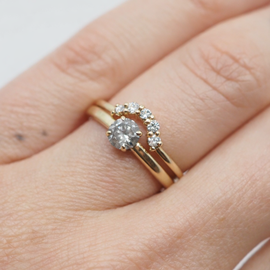Two gold rings on a person's finger against a light background. One ring with a diamond and the other ring in a curved shape with 5 white diamonds.