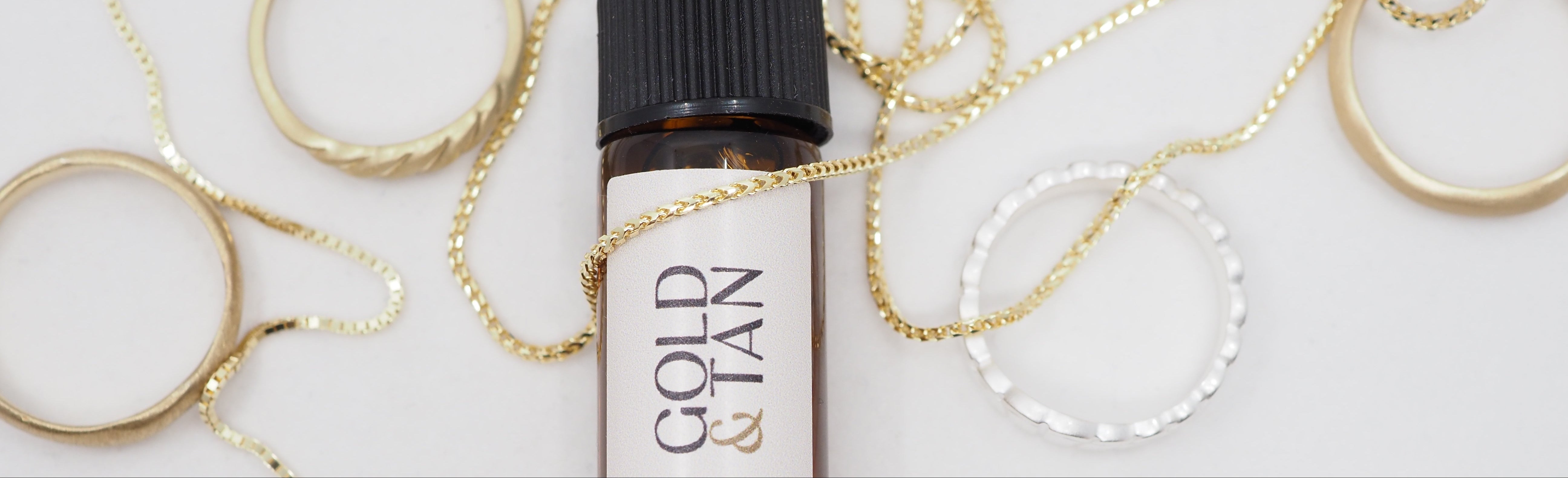 Small bottle of Gold & Tan body and bath oil surrounded by gold jewelry on a white background