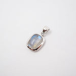 A close-up of a person wearing a delicate silver chain necklace with a small rectangular shaped pendant featuring a light blue-gray stone, styled with a dark brown knit cardigan.