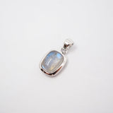 A close-up of a person wearing a delicate silver chain necklace with a small rectangular shaped pendant featuring a light blue-gray stone, styled with a dark brown knit cardigan.
