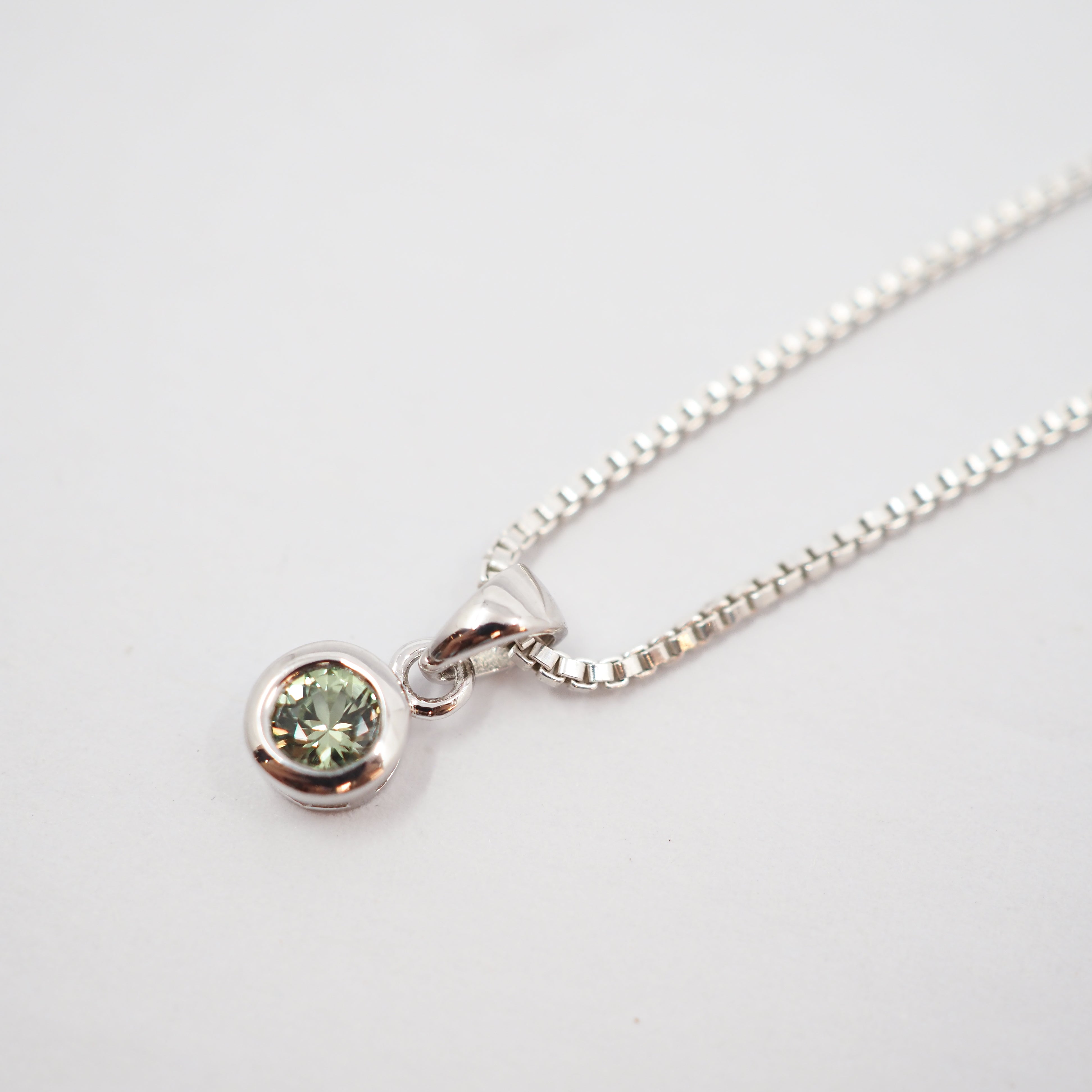 A silver box chain necklace with a small bezel-set round green gemstone pendant, photographed on a clean white background.