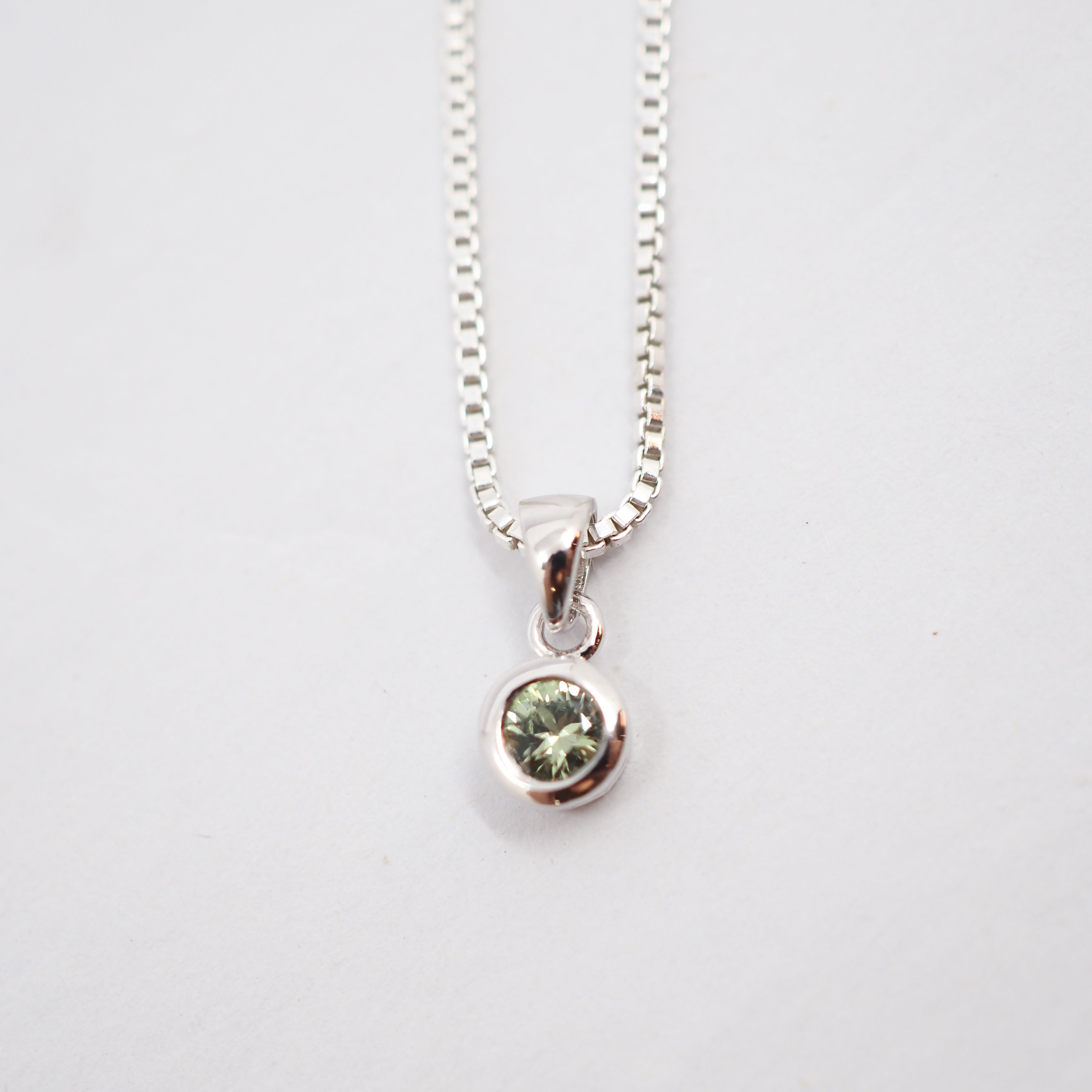 A silver box chain necklace with a small round bezel-set green gemstone pendant, shown against a plain white background.