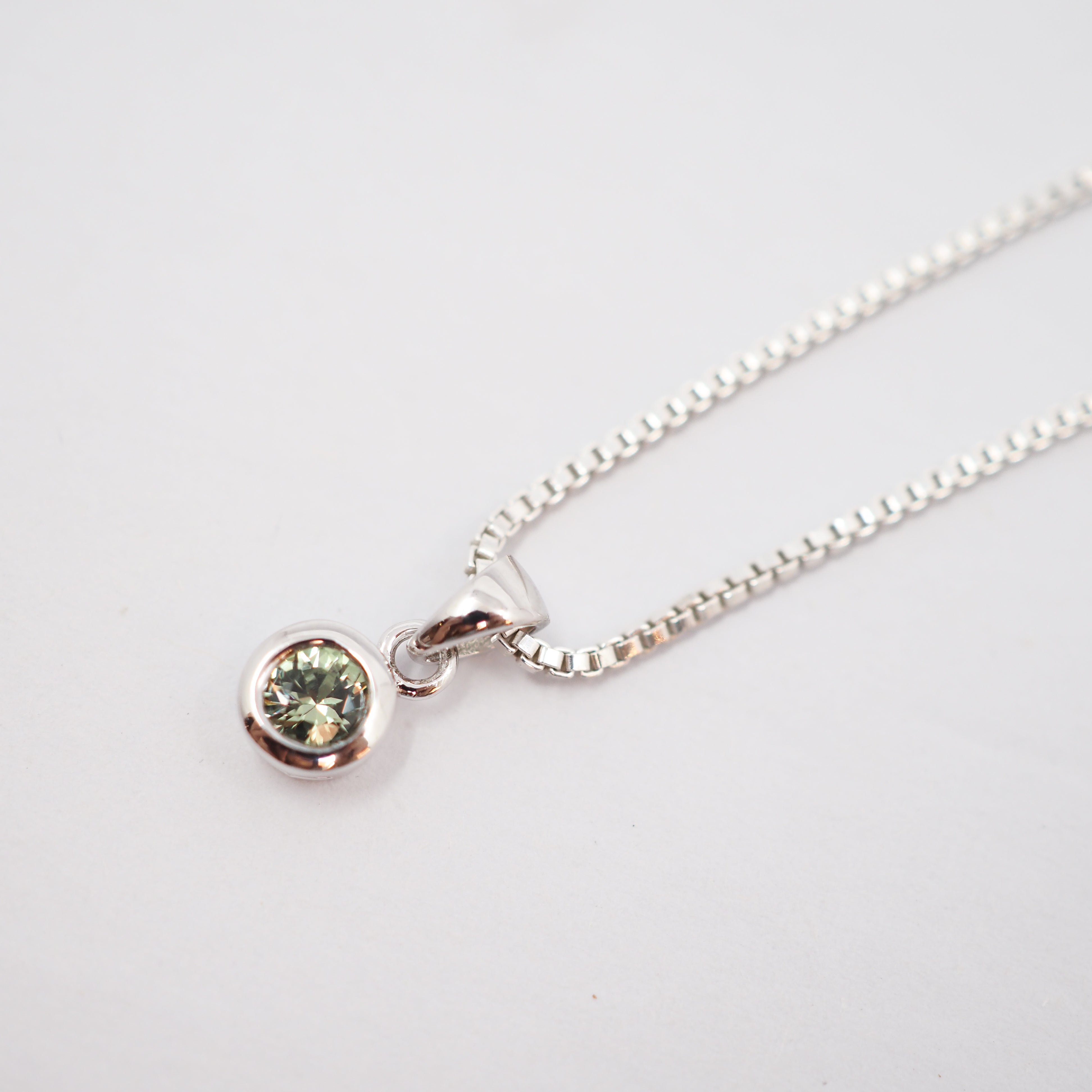 A silver box chain necklace with a small round bezel-set green gemstone pendant, shown on a plain white background.