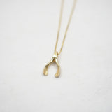 A delicate gold necklace with a small, wishbone pendant, photographed on a light gray textured background.