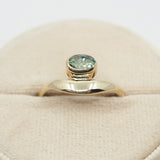 Gold ring with a green gemstone on a beige cushion against a white background
