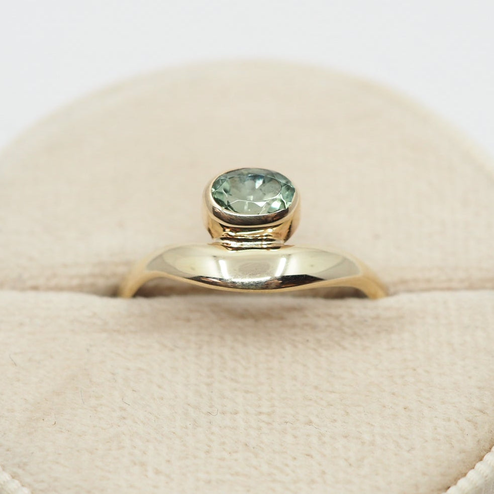 Gold ring with a green gemstone on a beige cushion against a white background