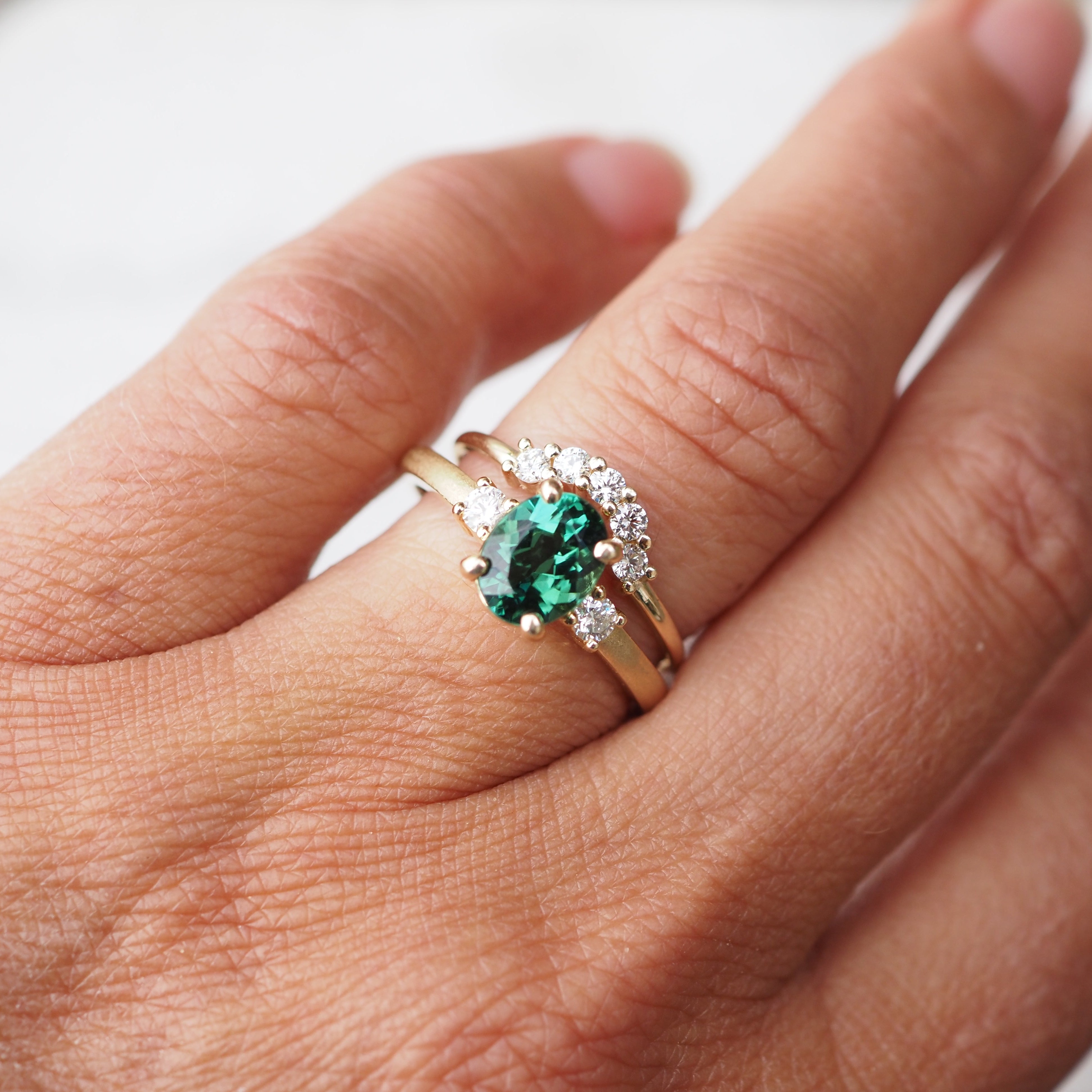 Hand wearing a ring with a green gemstone and curved diamond band on a neutral background