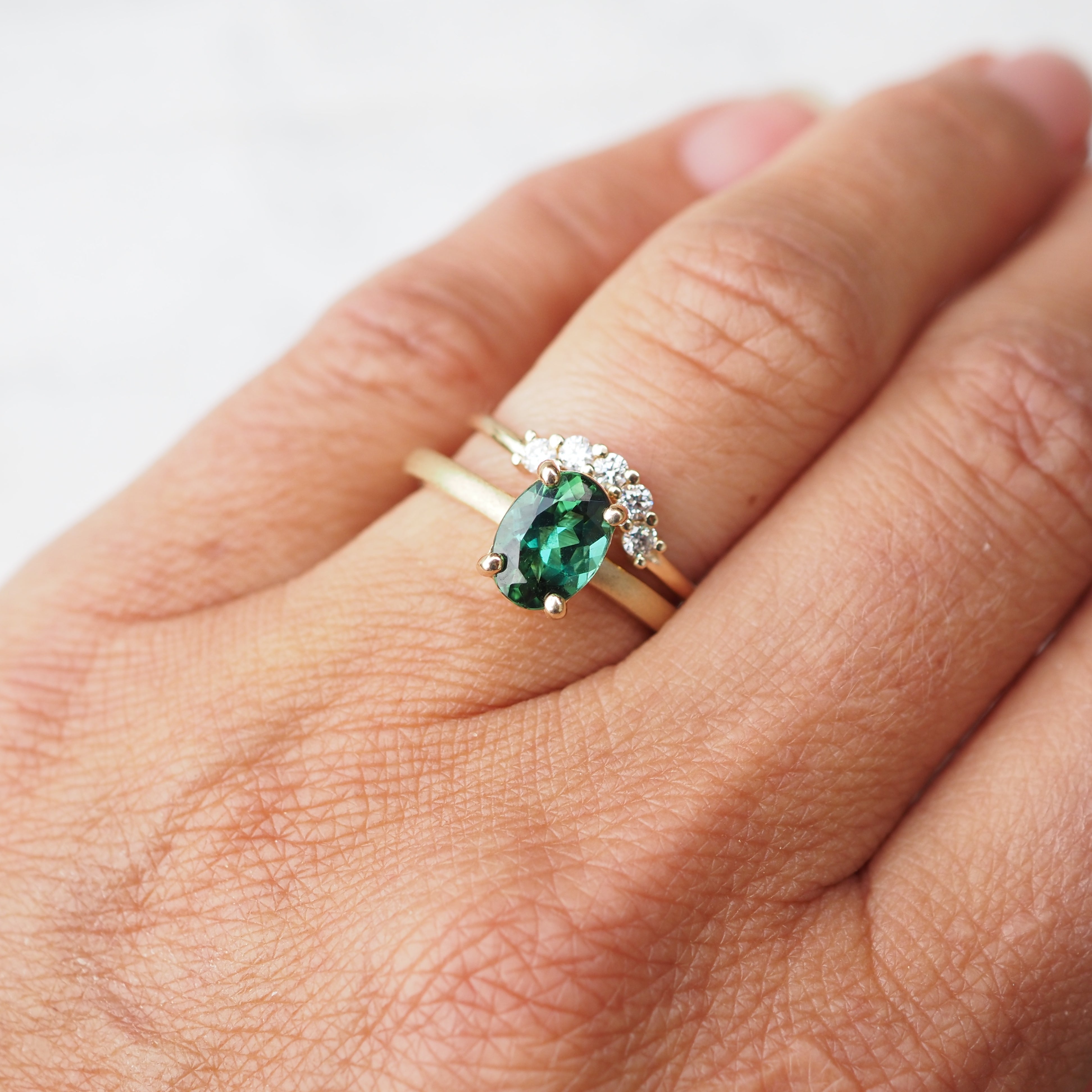 Hand wearing a gold ring with a green gemstone ring and a curved diamond band on a white background