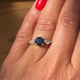 Ring with a blue gemstone and diamonds on a finger with red nail polish.