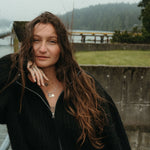 Woman with long hair standing outdoors near a body of water on a foggy day, wearing a jumble of pendants featuring the heart charm, the wave love charm, and the moyeha charm. 