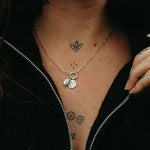 Person wearing a silver pendant jumble with a moyeha and lupine pendants, visible tattoos on neck and chest, against a dark background.