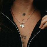 Person wearing a silver pendant jumble with a moyeha and lupine pendants, visible tattoos on neck and chest, against a dark background.
