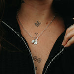 Person wearing a silver necklace with two round pendants, visible tattoos on neck and chest, against a dark background.