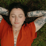 Person lying on grass with arms above head wearing an orange top