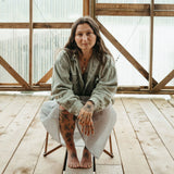 Woman sitting on a wooden chair in a rustic indoor setting with large windows, wearing the moyeha pendant. 