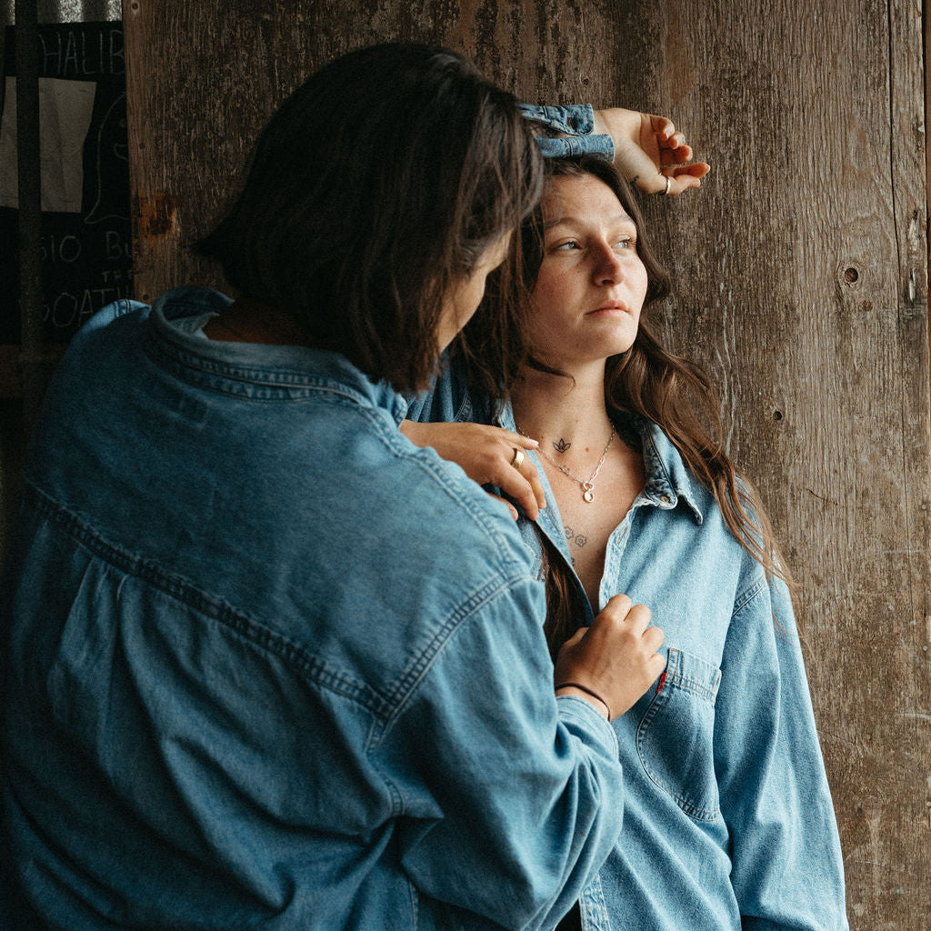 Two women wearing denim shirts standing close to each other against a wooden wall.