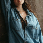 Person wearing a light blue denim shirt against a wooden background wearing the  lupine pendant. 
